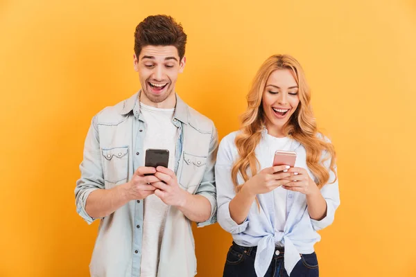 Photo of excited people man and woman smiling while both using cell phones isolated over yellow background