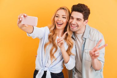 Genç adam ve kadın selfie cep telefonunda kamera zafer işareti üzerinde sarı arka plan izole el hareketi sırasında fotoğrafta portre