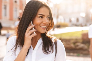Smartphone ile konuşmak ve uzak açık havada seyir gömlekli gülen esmer kadın resim