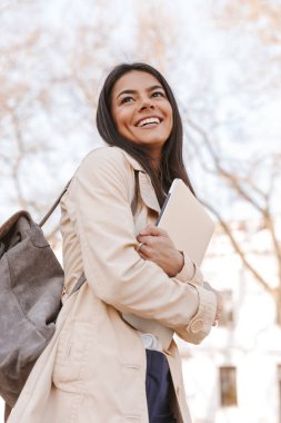 Bir şehir sokak üzerinde yürürken dizüstü taşıyan sonbahar ceket giymiş genç kadın gülüyor