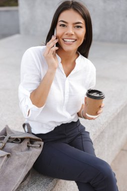 Açık havada süre smartphone ile konuşmak ve kameraya bakarak fincan kahve ile oturan gülen iş kadın dikey görüntü