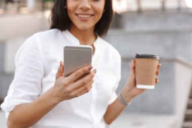Cep telefonunuzun ve açık havada take away kahve yudumlarken bir gülümseyen kadın resim kırpılmış