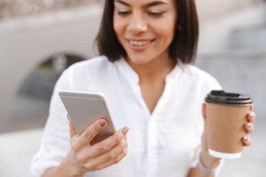 Memnun bir kadının açık havada take away kahve yudumlarken cep telefonuyla görüntü kırpılmış