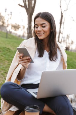 Cep telefonu ile dizüstü bilgisayar açık havada bir bankta otururken tutan genç ve güzel kadın