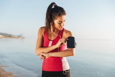 Genç güzel spor kadın smartphone kullanarak ve deniz açık havada yakınındaki müzik dinleme sırasında çalıştırmak için hazırlanma