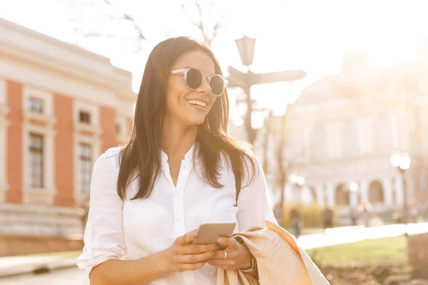 Mulher Morena Sorridente Camisa Óculos Sol Segurando Smartphone Olhando Para — Fotografia de Stock