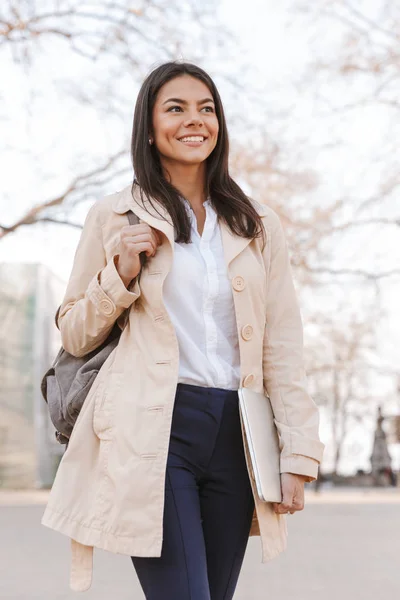 幸せな若い女は 街の通りを歩きながらノート パソコンを運ぶ秋のコートに身を包んだ — ストック写真