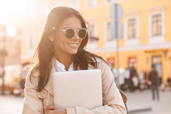 微笑的黑发妇女在夹克和太阳镜拥抱笔记本电脑 而看着户外 — 图库照片