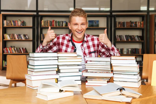 Sorrindo Adolescente Sentado Mesa Biblioteca Com Grandes Pilhas Livros Mostrando — Fotografia de Stock