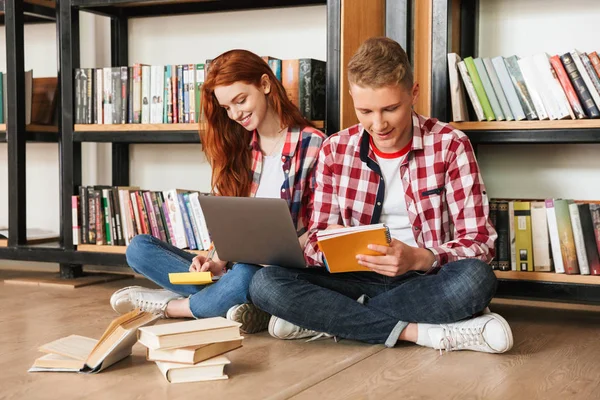 代のカップル図書室の本棚で床に座って ノート パソコンと宿題を笑みを浮かべてください — ストック写真