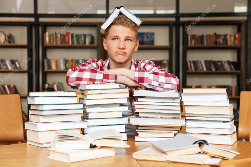 Upset teenage boy sitting at the library table with big stacks of books