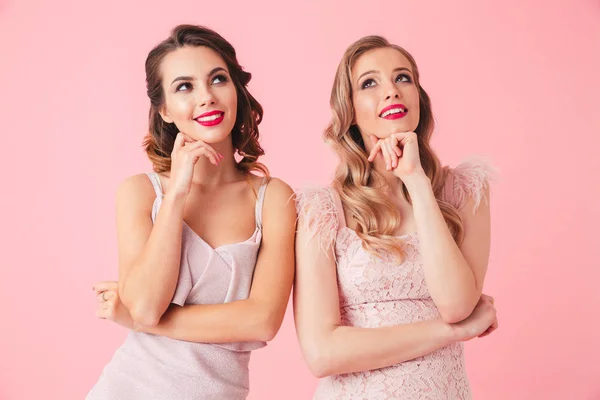 两个沉思的微笑妇女在礼服抚摸着她的下巴 并期待在粉红色背景 — 图库照片