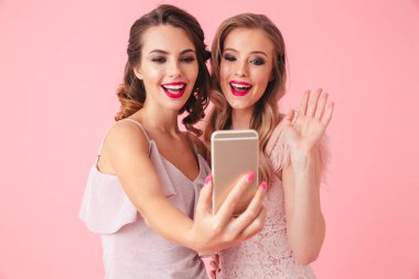 Fotoğraf gülümseyen ve cep telefonu pembe arka plan izole selfie fotoğraf çekerken fotoğraf makinesi vasıl el sallayarak iki genç parti kadın