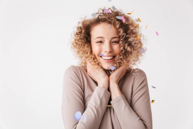 Portrait of a cheerful young curly blonde girl in dress playing with colorful confetti isolated over white background