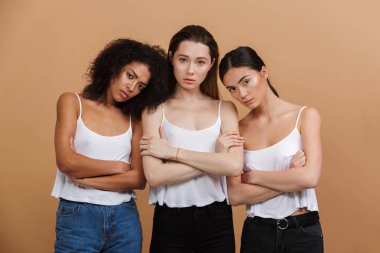 Üç ciddi Interracial kadın görüntüsünü: beyaz, Asya ve Afrika kökenli Amerikalı kızlar silah ile ayakta kot pantolon bej renkli arka plan üzerinde izole geçti