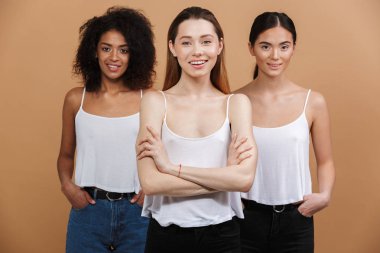 Görüntü farklı milletin üç mutlu gülümseyen kadınların: birlikte izole bej renkli arka plan üzerinde beyaz, Asya ve Afrika kökenli Amerikalı kız ayakta kot pantolon