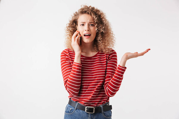 Портрет смущенной молодой девушки с кудрявыми волосами, разговаривающей по мобильному телефону на белом фоне
