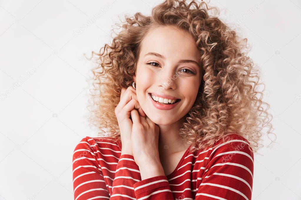 Smiling blonde curly woman in casual clothes posing with arms near face and looking at the camera over grey background