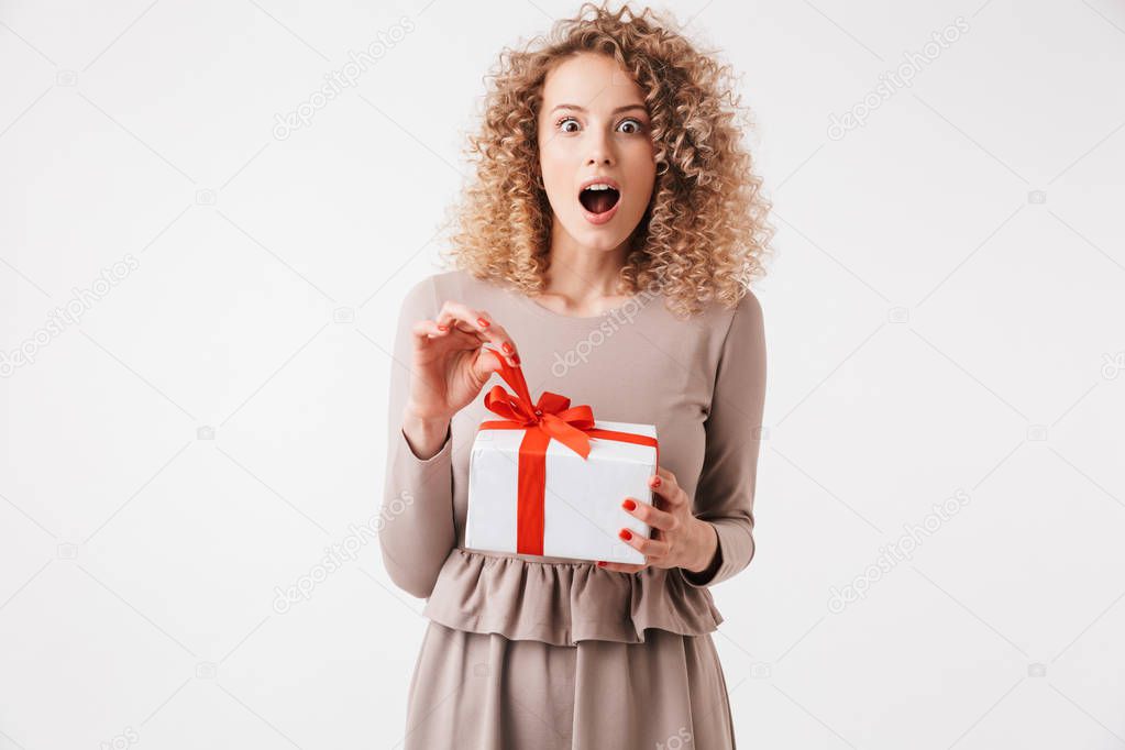 Portrait of an excited young curly blonde girl in dress holding present box isolated over white background