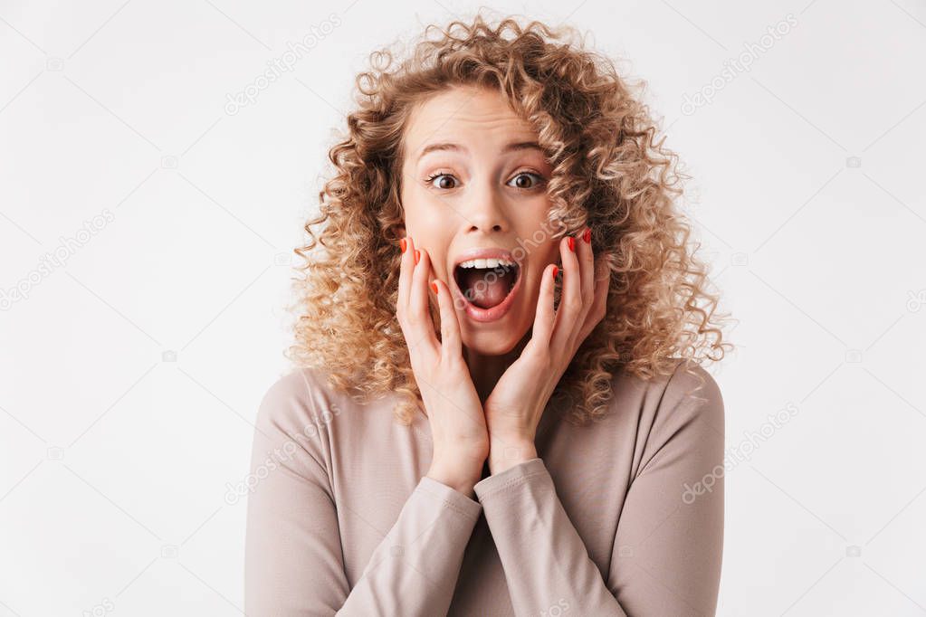 Surprised happy blonde curly woman in dress touching her cheeks and looking at the camera over grey background