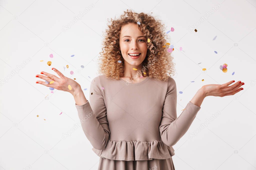 Portrait of a smiling young curly blonde girl in dress playing with colorful confetti isolated over white background