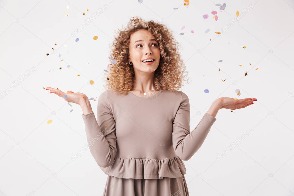 Portrait of a happy young curly blonde girl in dress playing with colorful confetti isolated over white background
