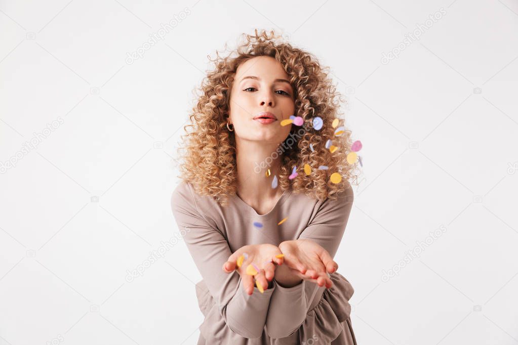 Portrait of a lovely young curly blonde girl in dress playing with colorful confetti isolated over white background