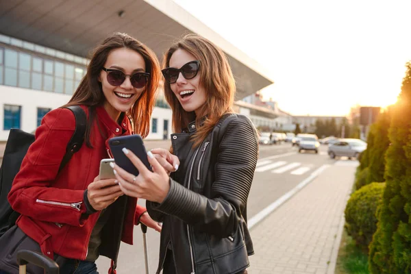 Bild Två Glada Europeiska Kvinnor Bär Solglasögon Tittar Smartphone Stående — Stockfoto