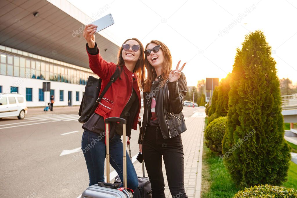 Image of two young caucasian women wearing sunglasses taking selfie photo on smartphone while standing with luggage near airport waiting for flight or after departure. Air travel concept