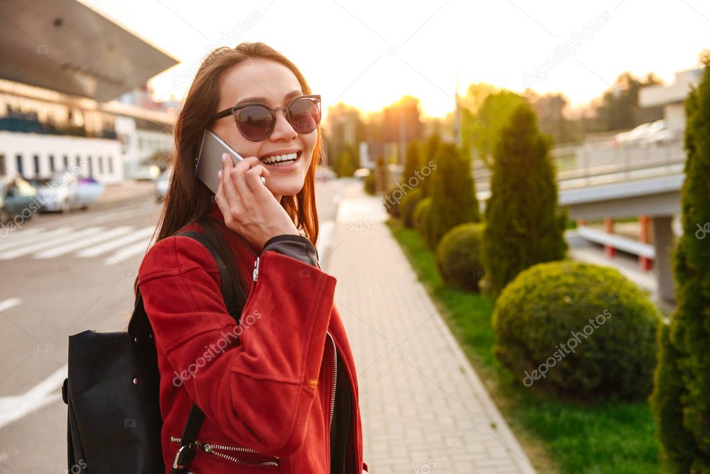 Portrait of happy young woman wearing sunglasses smiling while standing near airport and speaking on smartphone. Air travel or vacation concept