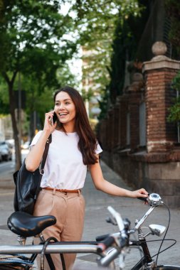 Görüntü şaşırtıcı gülümseyen güzel kadının açık havada cep telefonuyla konuşurken bisiklet üzerinde yürüyen.