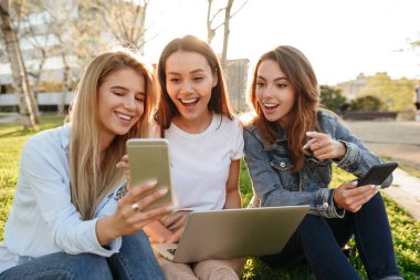 Park açık havada bir kenara dizüstü bilgisayar ve cep telefonu kullanarak bakarak çimenlerin üzerinde oturan inanılmaz üç arkadaş kadın fotoğrafı.