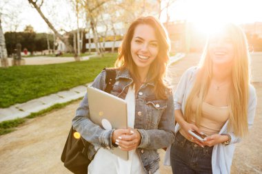 Park açık havada tutarak dizüstü bilgisayar ve cep telefonu kamera seyir mutlu iki arkadaş kadınların görüntü.