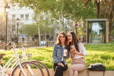 Görüntü açık havada birbirleri ile kahve yeme kruvasan içme konuşurken parkta oturan iki genç güzel kadın arkadaş.