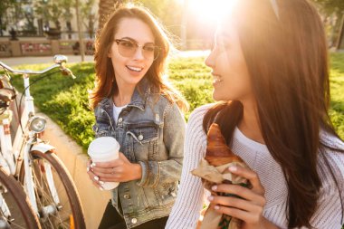 İki genç güzel kadın arkadaşlar açık havada Bisiklet Park birbirleri ile kahve yeme kruvasan içme söz ile görüntü.