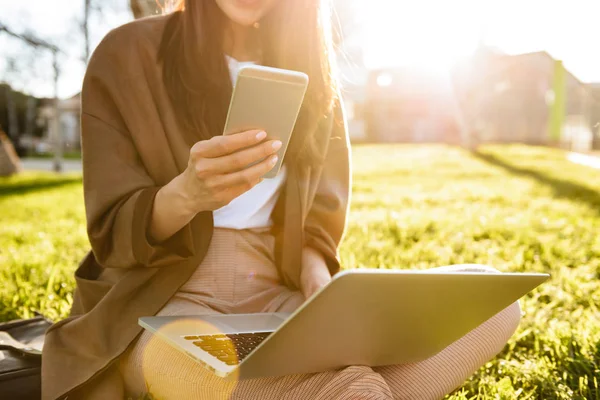 ラップトップ コンピューターや携帯電話を使用した屋外の芝生の上に座っている女性の写真をトリミング — ストック写真