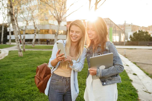 Image of happy two friends women in park outdoors holding laptop computer and using mobile phone talking with each other.