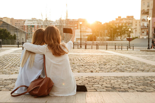 Back view photo of two friends women outdoors sitting and hugging while take a selfie.