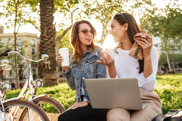 两个年轻快乐的女性朋友的照片户外与自行车公园喝咖啡吃牛角面包 而使用笔记本电脑 — 图库照片