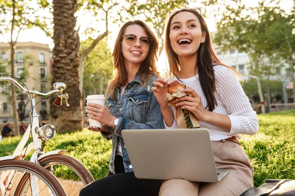 两个年轻快乐的女性朋友的照片户外与自行车公园喝咖啡吃牛角面包 而使用笔记本电脑 — 图库照片