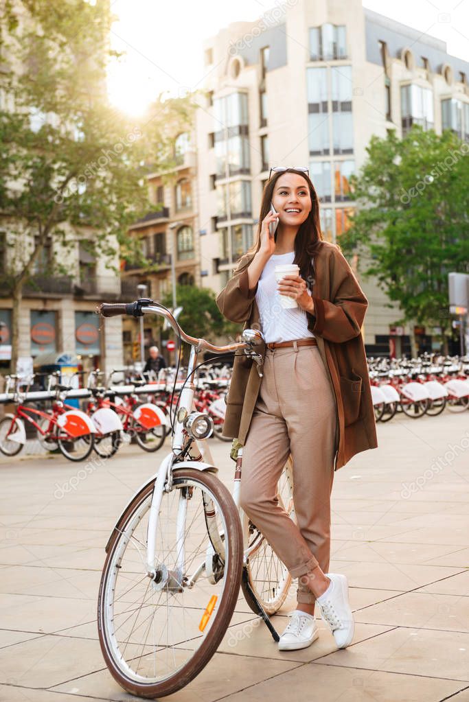 Photo of young pretty woman outdoors with bicycle on the street looking aside talking by mobile phone drinking coffee.