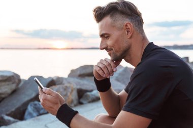 Plajda otururken cep telefonuyla gülümseyen bir sporcu portresi