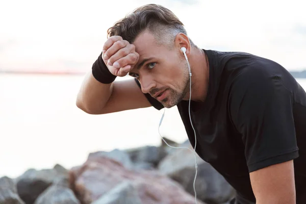 Close up portrait of a exhausted sportsman listening to music with earphones while resting after jogging at the beach
