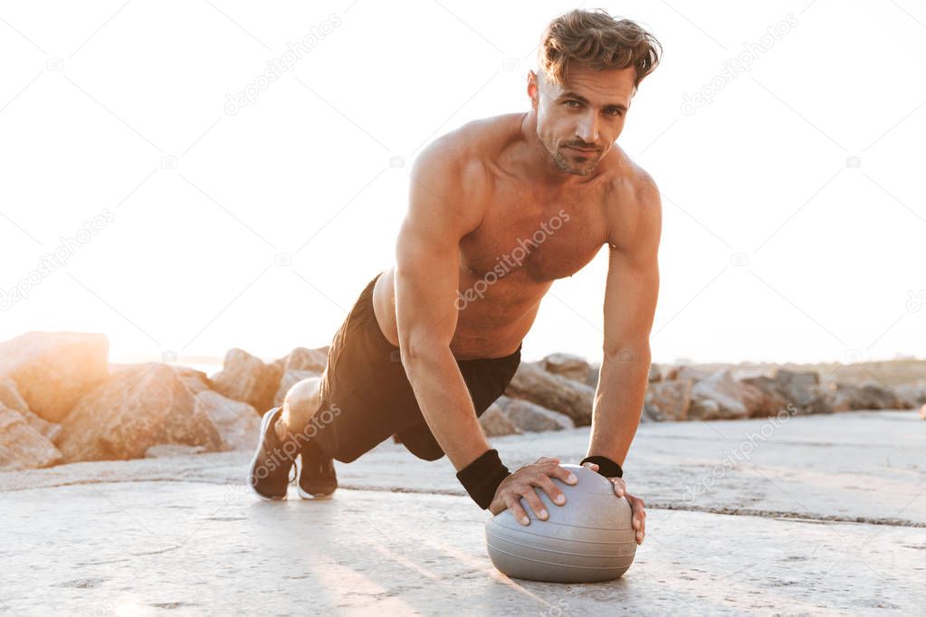 Portrait of a handsome shirtless sportsman doing exercises with small fitness ball outdoors