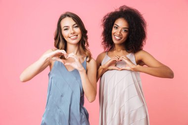 İki neşeli genç kız ile parmak pembe arka plan üzerinde izole kalp jest gösterilen yaz elbise giymiş