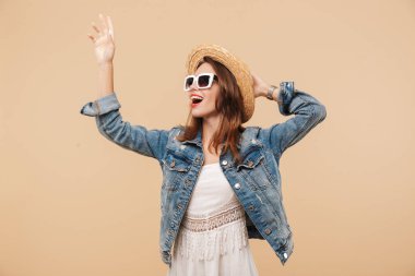 Yazlık giysiler ve güneş gözlüğü ile bej renkli arka plan üzerinde izole el sallayarak gülümseyen genç bir kız portresi
