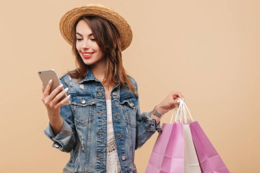 Alışveriş torbaları bej renkli arka plan üzerinde izole tutarken cep telefonunuzun yaz giysileri gülümseyen genç bir kız portresi