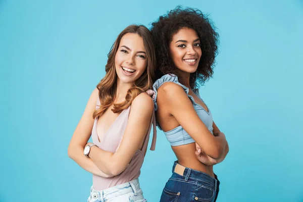 Two smiling young girls dressed in summer clothes standing back to back and looking at camera isolated over blue background