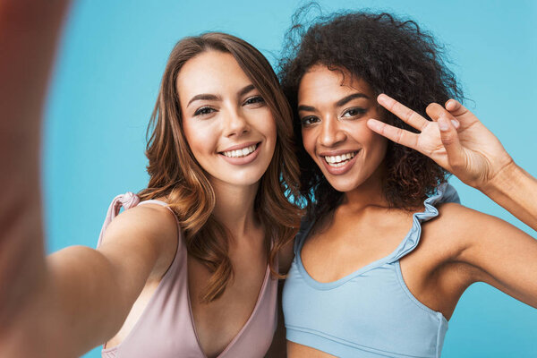 Two smiling young girls dressed in summer clothes taking a selfie isolated over blue background