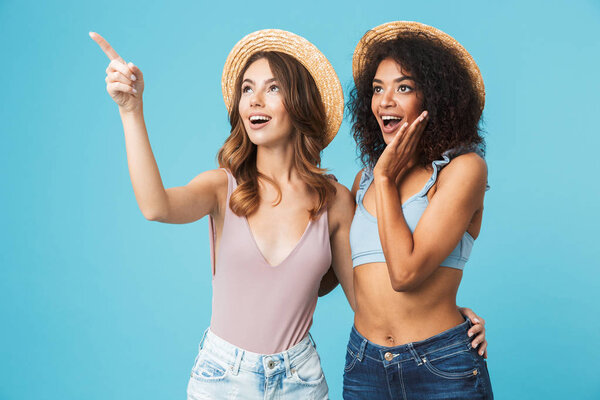 Two excited young girls dressed in summer clothes pointing at copy space isolated over blue background